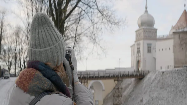 Una mujer en ropa de abrigo tomando fotos de la hermosa arquitectura de invierno de la ciudad vieja en una cámara. Acción. Concepto de turismo y viajes. — Foto de Stock