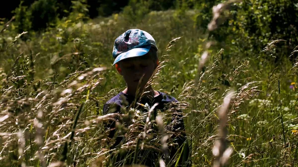 一个小机器人站在那里，眼睛紧闭在长草中的画像。创意。一个男孩在夏天的草地上，草地在风中摇曳. — 图库照片