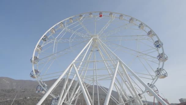 Vista inferior da roda gigante branca. Acção. Grande roda gigante no fundo do céu azul. Parque de diversões com roda gigante no fundo das montanhas no dia ensolarado — Vídeo de Stock