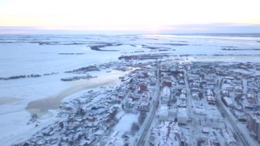 Drone 'dan kış görüntüsü. Clip. Tüm evlerin karla kaplı olduğu yüksek bir yerden şehrin manzarası, arabalı yollar, donmuş bir nehir ve birkaç ağaç ve bulutsuz parlak bir gökyüzü görebilirsiniz..