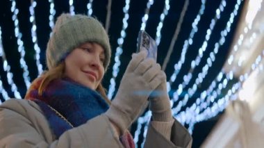Şehir caddesinde Noel aydınlatmalı bir kadın. Başla. Sıcak elbiseli bir kadın akıllı telefonuyla fotoğraf ya da video çekiyor, gülümsüyor..