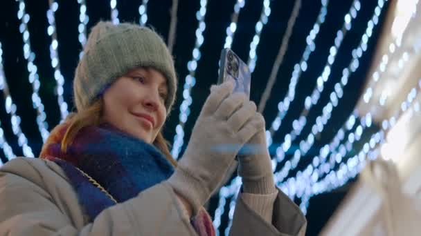 Mujer en la calle de la ciudad con iluminación navideña. Acción. Mujer en ropa de abrigo tomando fotos o videos en su teléfono inteligente sonriendo. — Vídeo de stock