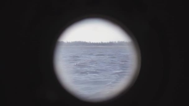 Pohled z kulatého okénka lodi na krajinu. CLIP. Pobřeží pokryté stromy, kterými loď pluje na klidném moři — Stock video