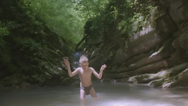 Dziecko w kąpielówkach bawi się i chlapie wodą stojącą w małym górskim stawie. KREATYWY. Blondyn lata i pływa w górskiej rzece — Wideo stockowe