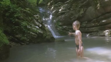 Çocuk şelalenin yanındaki gölde banyo yapıyor. - Yaratıcı. Bir çocuk neşeyle bir dağ şelalesine sıçrıyor. Adam parmağıyla işaret ediyor.