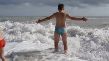 Çocuk kıyı boyunca yürür ve denize girer. - Yaratıcı. Mavi şortlu bir çocuk küçük dalgalarla denizde yüzmeye gider.