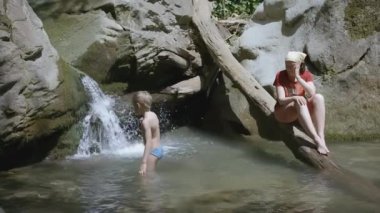 Sarı saçlı bir çocuk şelaleye yaklaşıyor. - Yaratıcı. Anne, oğlunun şelalenin yakınındaki küçük bir dağ nehrinde yüzüşünü izliyor.