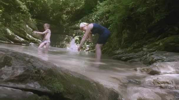 Mamma si lava le mani in un piccolo stagno di montagna mentre suo figlio spruzza. Creativo. Un ragazzo biondo si gode l'estate e schizza in acqua — Video Stock