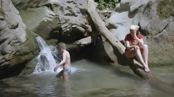 Un ragazzo con i capelli biondi si avvicina a una cascata su un piccolo fiume di montagna. Creativo. Mamma si siede su un tronco e guarda suo figlio fare il bagno — Video Stock