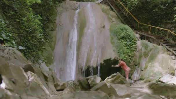 Eine Frau steht inmitten eines kleinen Flusses und bewundert den Wasserfall. CREATIVE. Eine Frau in rotem T-Shirt und kurzer Hose ging von der Touristenroute weg, um sich den Gebirgsfluss anzusehen — Stockvideo