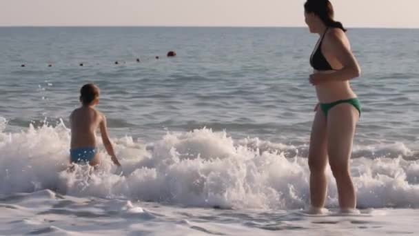 Matka obserwuje syna stojącego w morzu, pluskającego się i cieszącego się surfingiem. KREATYWY. Rodzina spędza letnie wakacje na wybrzeżu. — Wideo stockowe