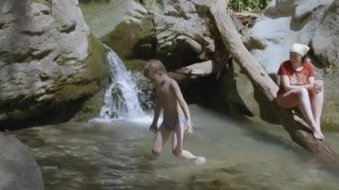 Çocuk bir şelalenin altındaki küçük bir gölden çıkar. - Yaratıcı. Anne, oğlunun şelalenin yakınındaki küçük bir dağ nehrinde yüzüşünü izliyor.