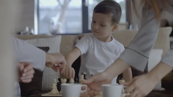 아이는 체스를 연주합니다. 행동이요. 어린 소년은 그의 부모와 체스를합니다. 집에서 시간을 보내는 가벼운 옷을 입은 가족 — 비디오