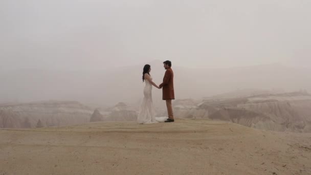 Schönes junges kaukasisches Brautpaar, Hochzeit in der Türkei, Kappadokien. Handeln. Luftaufnahme eines liebenden Paares, das Händchen haltend auf einem Gipfel eines Berges in der Nähe der Klippe steht. — Stockvideo