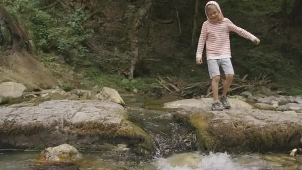 Piękny krajobraz z wodospadem.Kreatywny. Mały chłopiec w bluzie biegnie i skacze przez strumień na skałach, a las jest widoczny za nim.. — Wideo stockowe