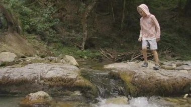 Şelalesi olan güzel bir manzara. Yaratıcı. Eşofmanlı küçük bir çocuk koşar ve kayalıklardaki dereden atlar ve orman arkada görünür..