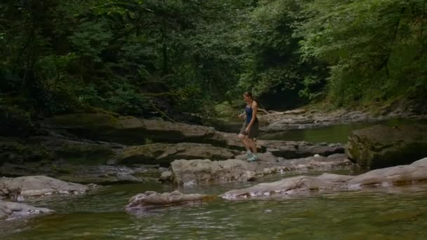 Una chica en pantalones cortos hinchados en la naturaleza.Creativa.Una mujer joven salta sobre piedra a través de un arroyo en la selva. — Vídeo de stock