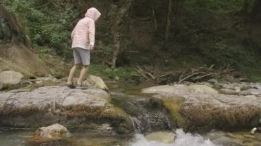 Dağlık su deresinin üzerinden atlayan bir çocuğun yan görüntüsü. Yaratıcı. Orman gölü ve bir çocuk eğleniyor..