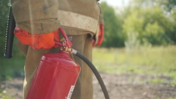 Στενή πλαϊνή όψη πυροσβέστη με κόκκινο πυροσβεστήρα σε στολή ασφαλείας σώματος για προστασία από πυρκαϊά. Κλιπ. Άνδρας πυροσβέστης στέκεται στο πράσινο πεδίο. — Αρχείο Βίντεο