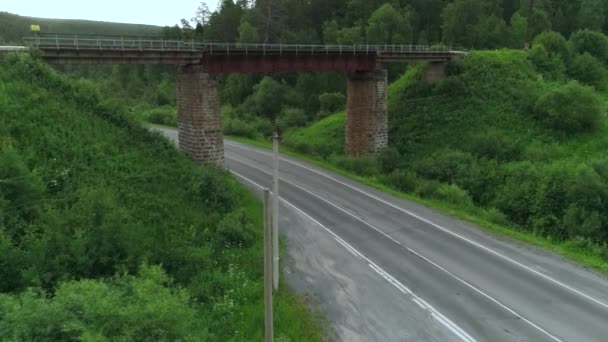 森の下に道路がある橋のトップビュー. シーンか。 森林地帯の田舎の高速道路を渡る古い橋. 夏の森林と丘陵地帯の道路を備えた橋 — ストック動画