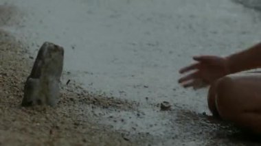 Kıyıda suyla oynayan çocuğun yakın çekimi. Yaratıcı. Kıyıda oturan çocuk su sıçratıyor. Çocuk dalgalarla su sıçratıyor.