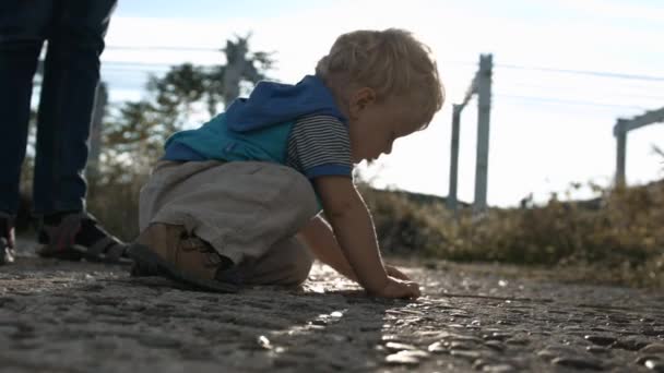 Oldalnézet egy gyermek játszik a kavicsos úton egy nyári napsütéses napon. Szőke fiú gyermek felfedező kövek az utcán, a szülei ott állnak a közelben. — Stock videók