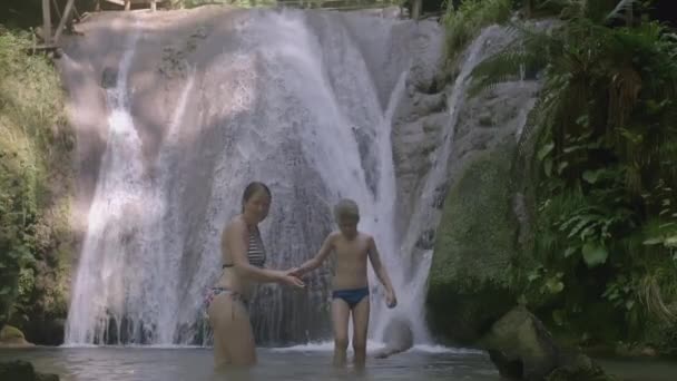 Una mujer le da su mano a un niño en el fondo de una cascada. Creativo. Madre e hijo están caminando a lo largo del estanque frente a la cascada. En la vegetación tropical es un lago con un — Vídeos de Stock