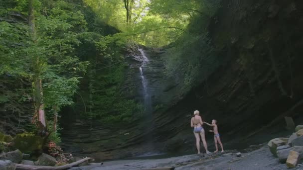 两个人站在瀑布的底部。恶心死了一个巨大的瀑布从山上掉下来.瀑布附近有两名游客。在丛林里，一条水流从高处落下 — 图库视频影像