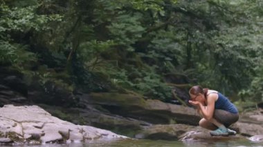 Kız gölden su içiyor ve avuçlarında topluyor. - Yaratıcı. Tişörtlü ve şortlu genç bir bayan göletin yanında oturdu. Ormanın ortasında, bir kız gölün kenarında duruyor.