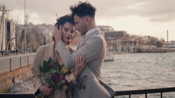 Hezký muž a krásná žena na rande u řeky stojící na mostě za větrného dne. Akce. Žena držící kytici květin a muž objímající ji. — Stock video