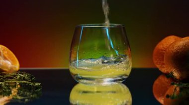 Masanın cam yüzeyindeki turunçgil kokteyli. Borsa klipsi. Meyveler ve biberiye yaprakları renkli ışıklandırılmış arka planda masanın üzerinde yatıyor.