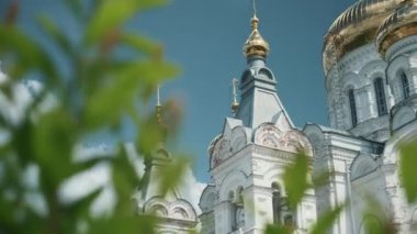 Ortodoks kilisesinin resimli görüntüsü. Şarjör. Beyaz kilisenin arka planında kubbeli yeşil dalların yakın çekimi. Yazın mavi gökyüzünün arka planında altın kubbeleri olan güzel beyaz kilise.. 