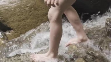 Bacakları kayalıklarda suyla kaplı, ağır çekimde. Yaratıcı. Dağlık bir derede, taşların üzerinde yürüyen genç bir çocuğun bacaklarını kapat.