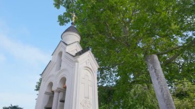 Kubbesinde altın haç olan beyaz bir kilisenin alçak açılı görüntüsü. Başla. Yeşil ağaçlı bir mimari bina. 