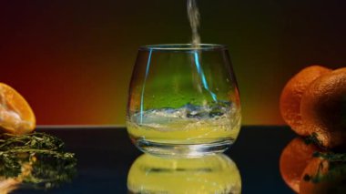 Masanın cam yüzeyindeki turunçgil kokteyli. Borsa klipsi. Meyveler ve biberiye yaprakları renkli ışıklandırılmış arka planda masanın üzerinde yatıyor.