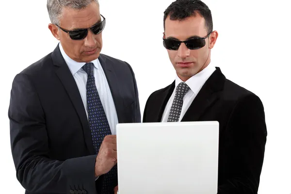 Hombres serios en trajes inteligentes con gafas de sol sosteniendo un ordenador portátil —  Fotos de Stock