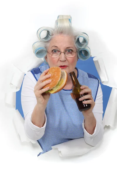 Elderly woman with her hair in rollers eating a hamburger and drinking beer — Stock Photo, Image