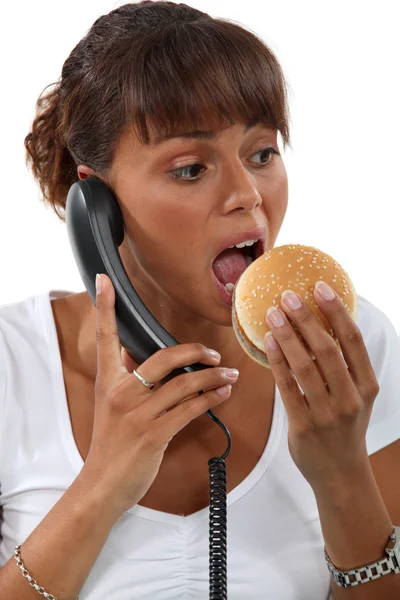 Mulher comendo um hambúrguer enquanto fala ao telefone — Fotografia de Stock