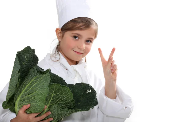 Uma menina vestida de uniforme de cozinheiro segurando um repolho e fazendo o sinal v — Fotografia de Stock