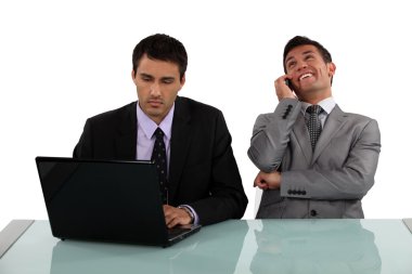 Serious businessman sitting next to a laughing colleague clipart