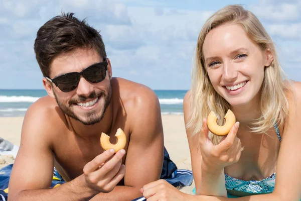 一对笑着的年轻夫妇在海滩上吃饼干 — 图库照片