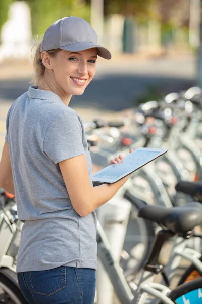 有平板电脑的妇女站在一排租来的自行车旁边 — 图库照片