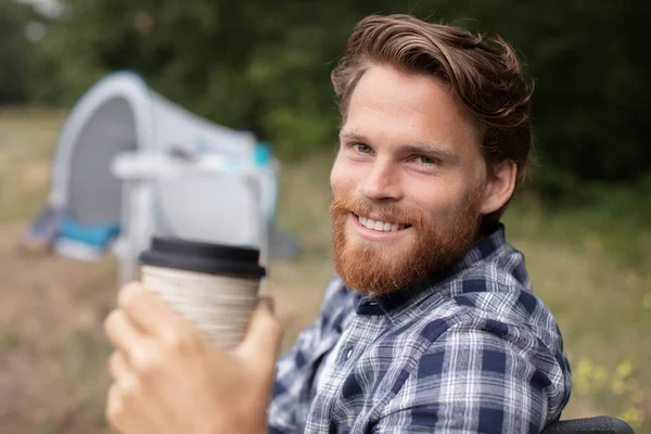 smiling young beared man drinking tea at camping