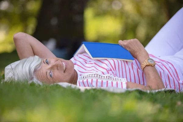 Starsza Kobieta Leży Zielonej Trawie Czyta Jej Książkę — Zdjęcie stockowe