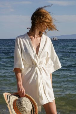 Plajda keten kimono elbise ve büyük hasır şapka giyen genç bir kadının portresi.