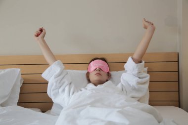 Pembe uyku maskeli genç, uykulu kadın sabahları yatakta geriniyor.