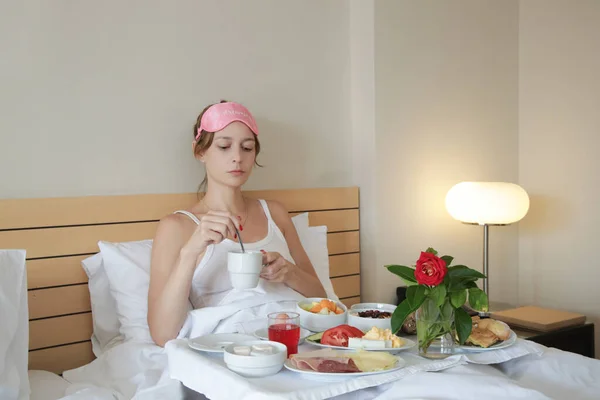 若いです眠いです女性とともにピンク睡眠マスク持っています朝食でザベッド — ストック写真