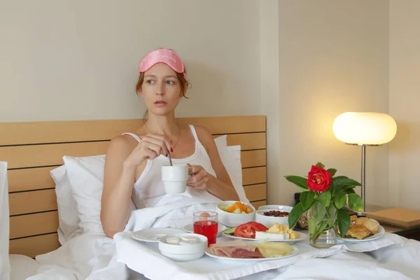 若いです眠いです女性とともにピンク睡眠マスク持っています朝食でザベッド — ストック写真