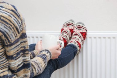 Elinde bir fincan sıcak içecek ve ayakları üzerinde renkli örgü çoraplar tutan kadınlar radyatör ısıtıcısında ısınıyor. Tanık zamanı, soğuk hava yaşam tarzı.