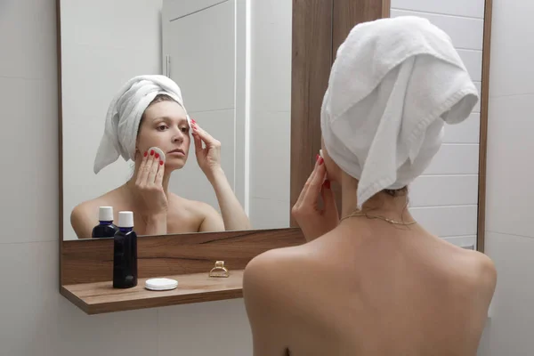 バスルームに立つ美しい若い女性の肖像画と鏡の前に綿のパッドで彼女の顔をきれいに 化粧を削除 美しさとスパのコンセプト — ストック写真
